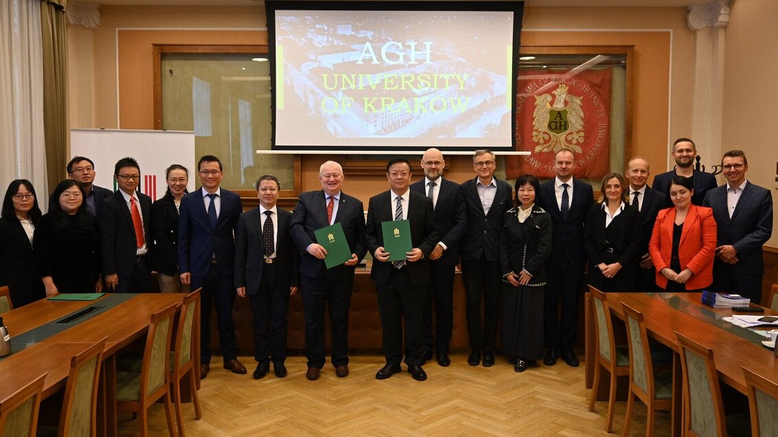 Image of a group of people in a large room posing for a photo, two men in the foreground are holding green folders; behind the group, there is a presentation screen with the name of the AGH University in large white letters