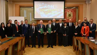 Image of a group of people in a large room posing for a photo, two men in the foreground are holding green folders; behind the group, there is a presentation screen with the name of the AGH University in large white letters