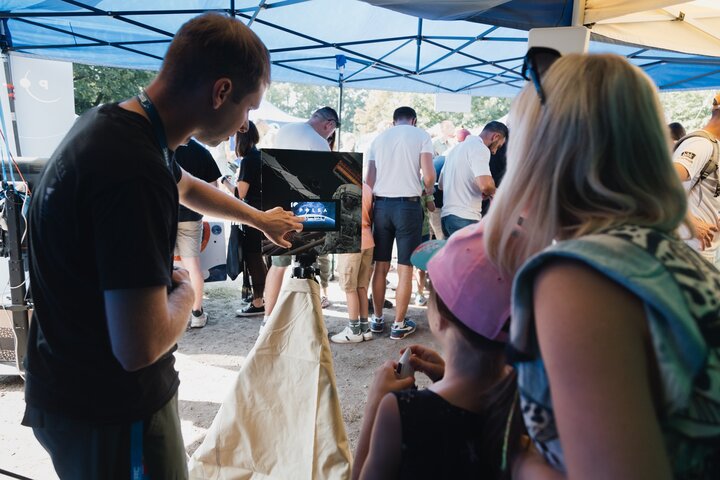 Image of a man showing a woman and a child an electronic device with a screen on a tripod, some people visible in the background