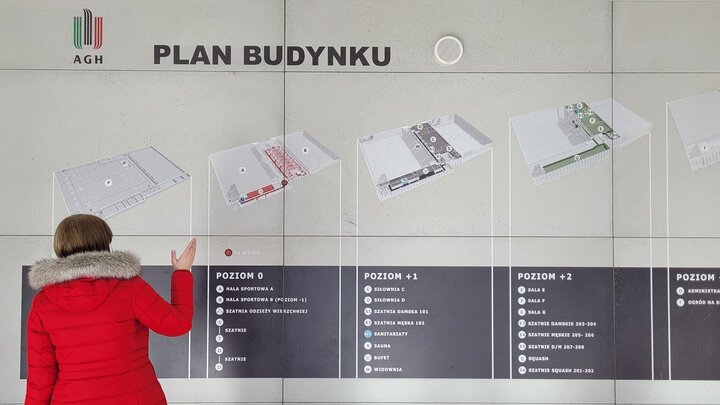 The image shows the plan of the building divided into floors. A woman in a red coat is pointing to the plan which is placed on a grey wall.