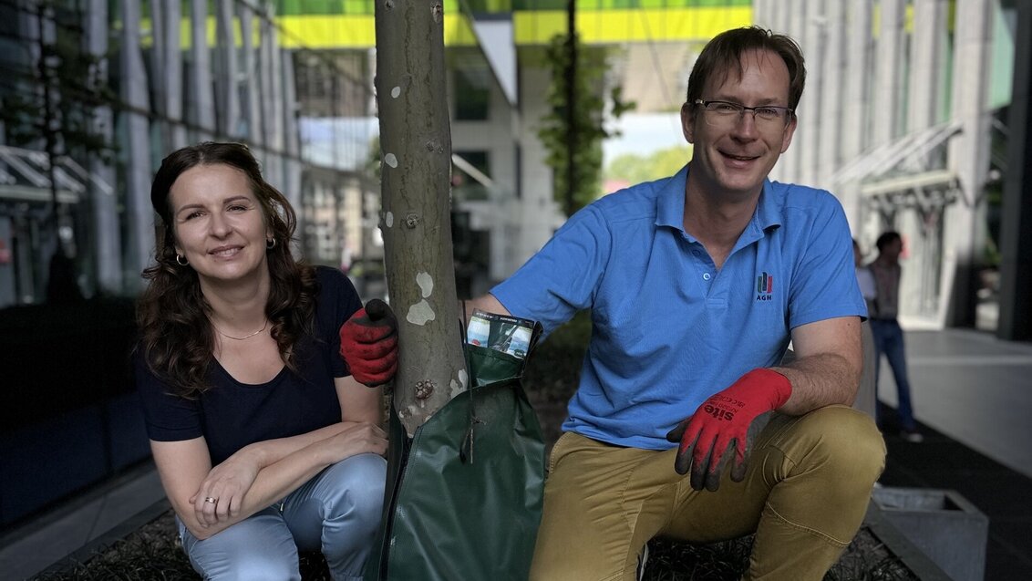 Image of a woman and a man smiling, squatting with a tree between the two of them, a modern grey and white building in the background