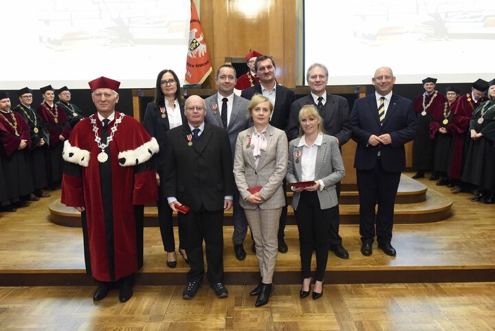 A formal group photo with people in business attire and a man in a red academic robe. They stand in front of a group of university authorities during an award ceremony in the formal AGH University Assembly Hall.