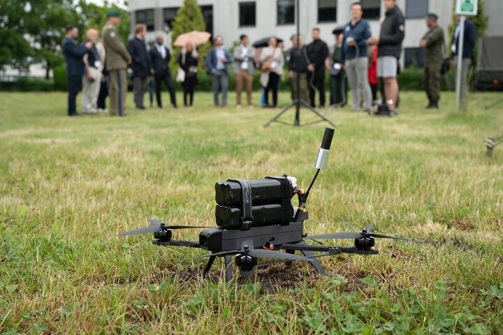 Image of a drone being tested on a field, a group of people visible in the background