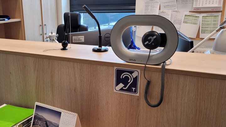 The image shows a portable induction loop device on a wooden reception desk, designed to assist people with hearing aids. A symbol with an ear and a "T" indicates telecoil compatibility, enhancing communication for individuals with hearing impairments.