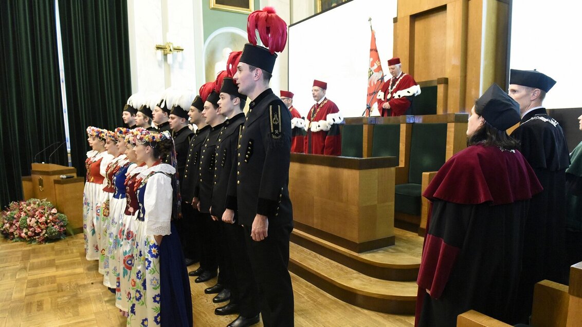 Image of a formal ceremony with students in traditional folk costumes and black mining uniforms standing in formation. Behind them, university authorities in red academic robes preside over the event from a podium in the mail assembly hall