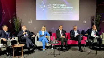 Image of a session at a congress, six people sitting at a stage, one man speaking to a microfone; behind them, there is a presentation screen with the name and the logo of the congress