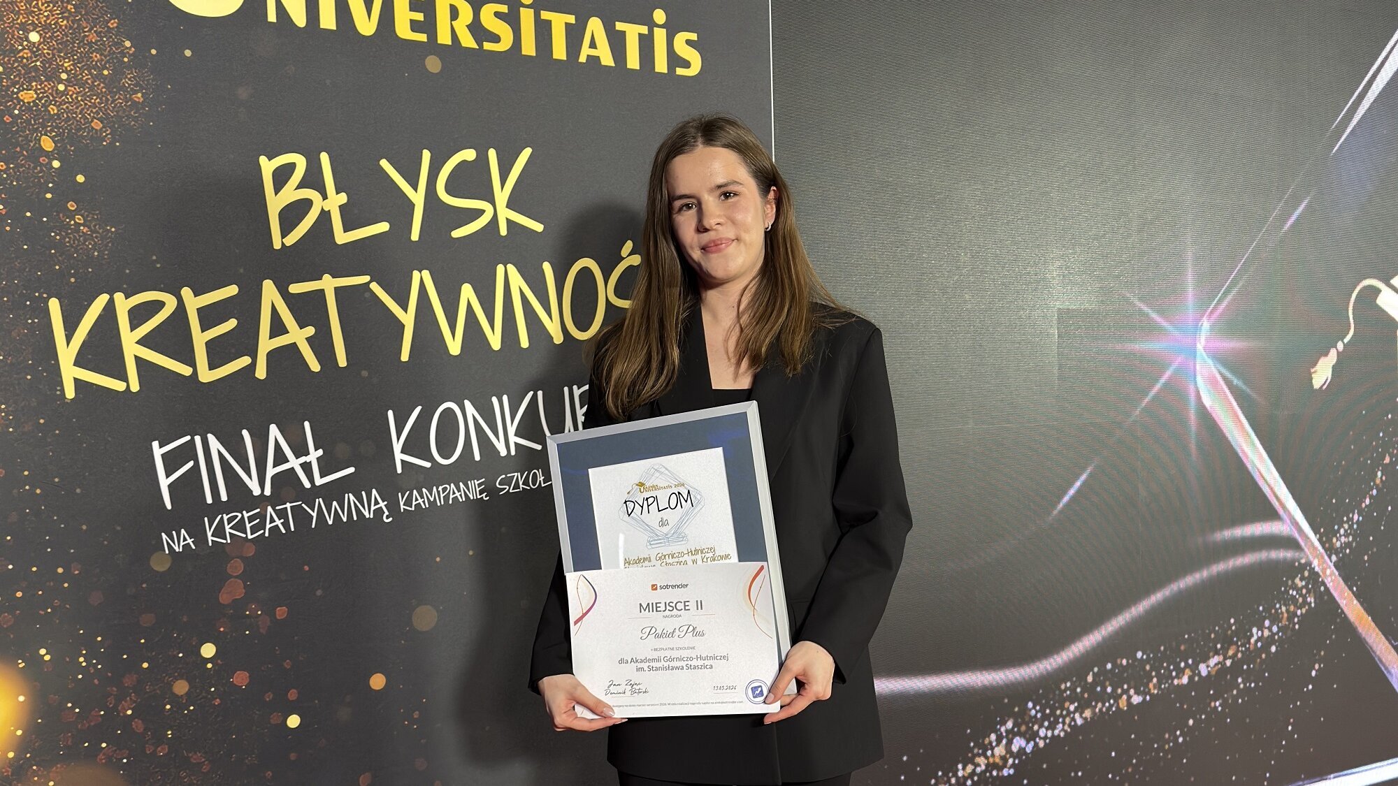 A young woman in a black suit posing against a dark-coloured competition backdrop holding a framed diploma.