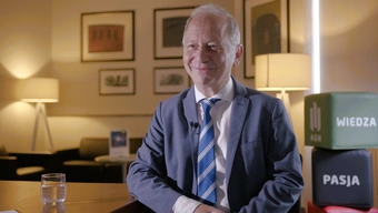 An image of an older man in a suit, a white shirt with a striped tie, smiling, seated at a table in a conference room with ambient lighting.